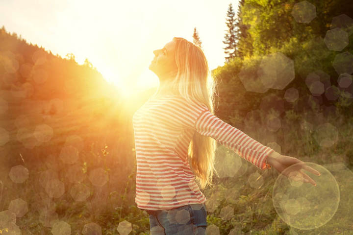 A beaufiul, confident, radiant woman with her arms spread wide looking up into the sky, with the sun behind her.