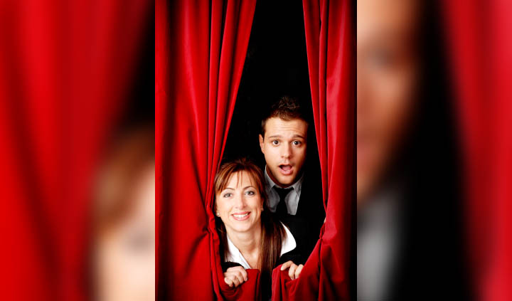 A man and a woman peek out from behind a curtain on a stage.