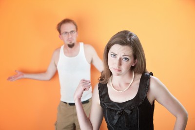 A beautiful woman is pointing at her slacker boyfriend wondering if she needs to move on. He is wearing a white tank top standing against an orange wall.