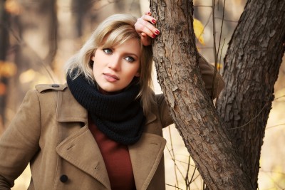 A beautiful blond woman leans against a tree during autumn, thinking why she stays in the relationship she is in.