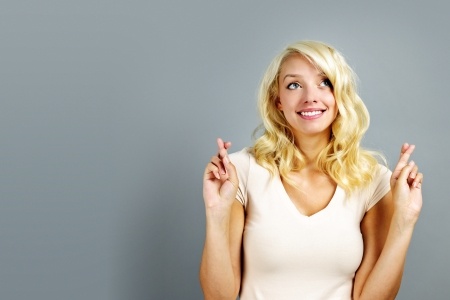 A beautiful blonde woman stands with her hands up and fingers crossed smiling and hoping to be chosen.