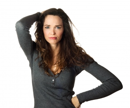 A beautiful brunette woman in a gray sweater has one hand on her head and one hand on her hip, sad and confused wondering if her expectations of her boyfriend are reasonable. 