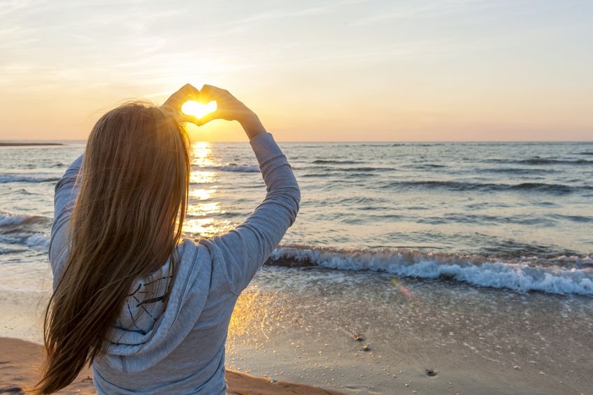 A beautiful woman holds her hands out in the shape of a heart while watching the sunset at the beach.