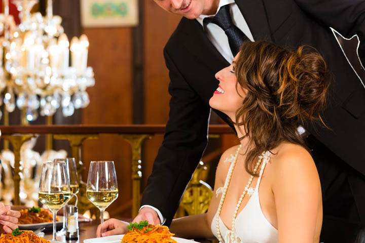 A beautiful, confident woman sits smiling at the table in a fine restaurant while the waiter delivers her food.