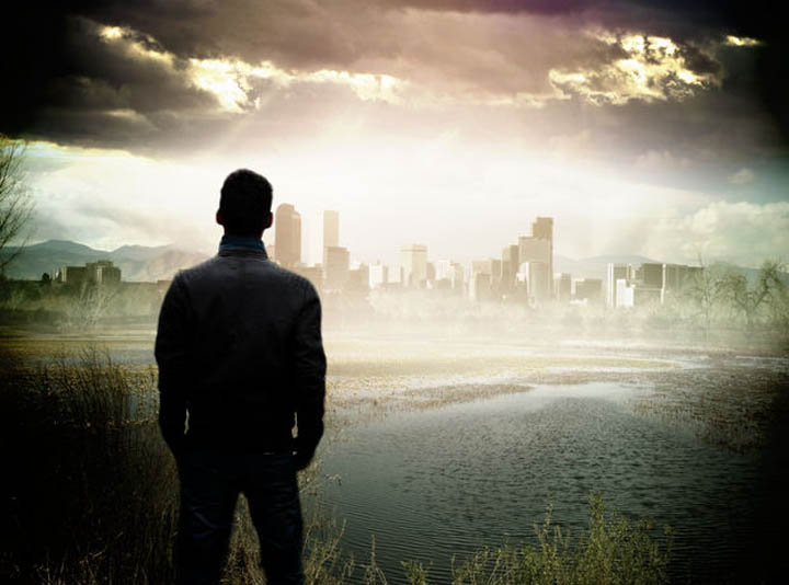 A man stands on the edge of a pond looking at the city in the distance.