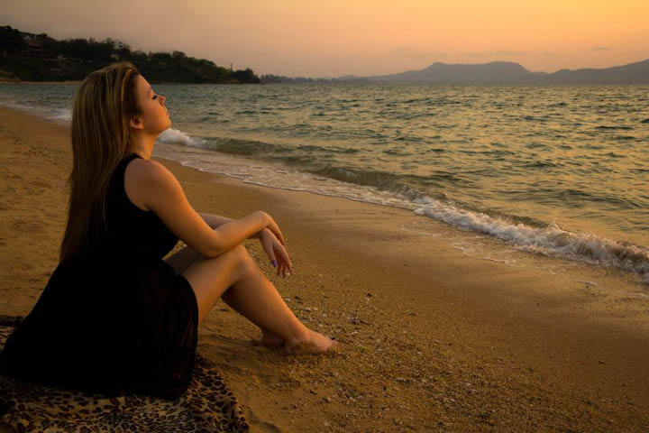 A beautiful woman is sitting on a beach looking at the ocean, wondering when he'll commit.