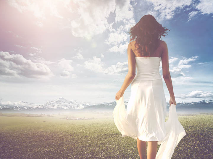 slender long-haired woman wearing white summer dress while walking on a green meadow towards a bright and sunny horizon.