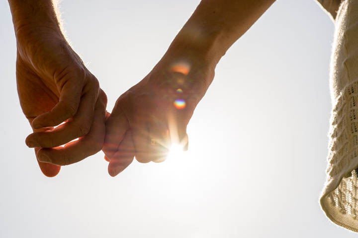 Close up of hands of romantic couple holding together loosely with sun rays on a white sky.