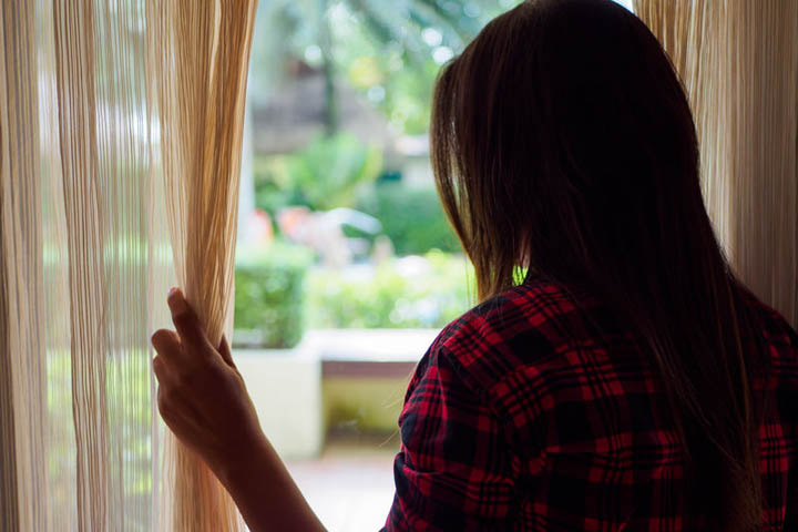 Rear view of a woman holding the curtains open to look out of a large light window at home.