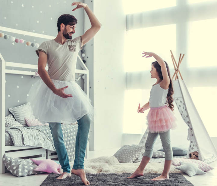 Cute little daughter and her handsome young dad in skirts are dancing and smiling while playing together in child's room