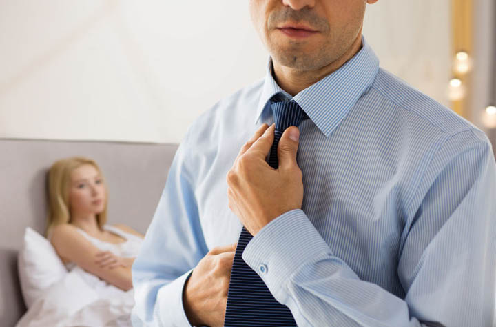 Relationship problems concept - close up of man in shirt dressing up and adjusting tie on neck over woman in bed background.