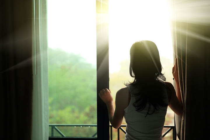 A beautiful woman looks out the window at the sun shining through realizing she has a fresh start.