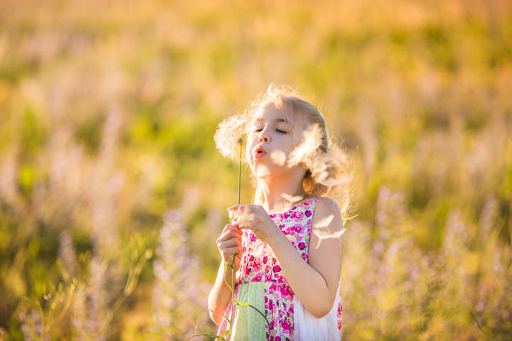 Beautiful little girl with big dreams, standing in a field blowing dandelion seeds.